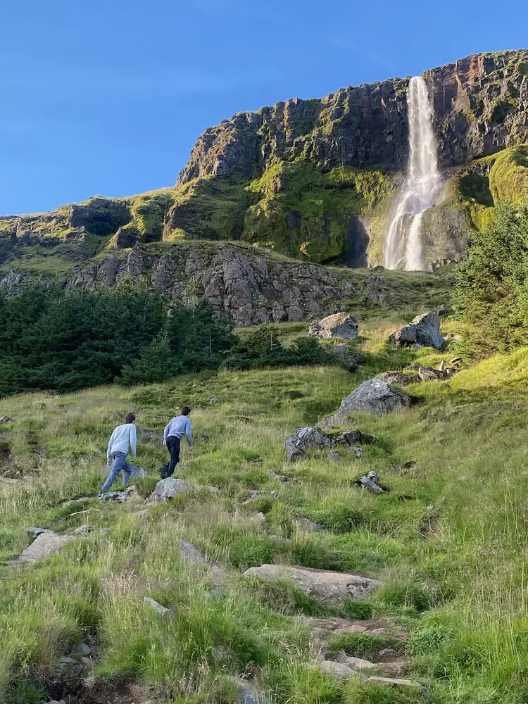 Two hikers climbing a grassy, boulder-strewn slope toward a tall waterfall pouring off a mossy cliff.